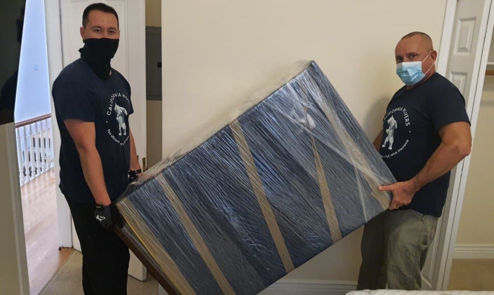 Two California Movers crew members carrying a shrink-wrapped mattress through a doorway during a residential move